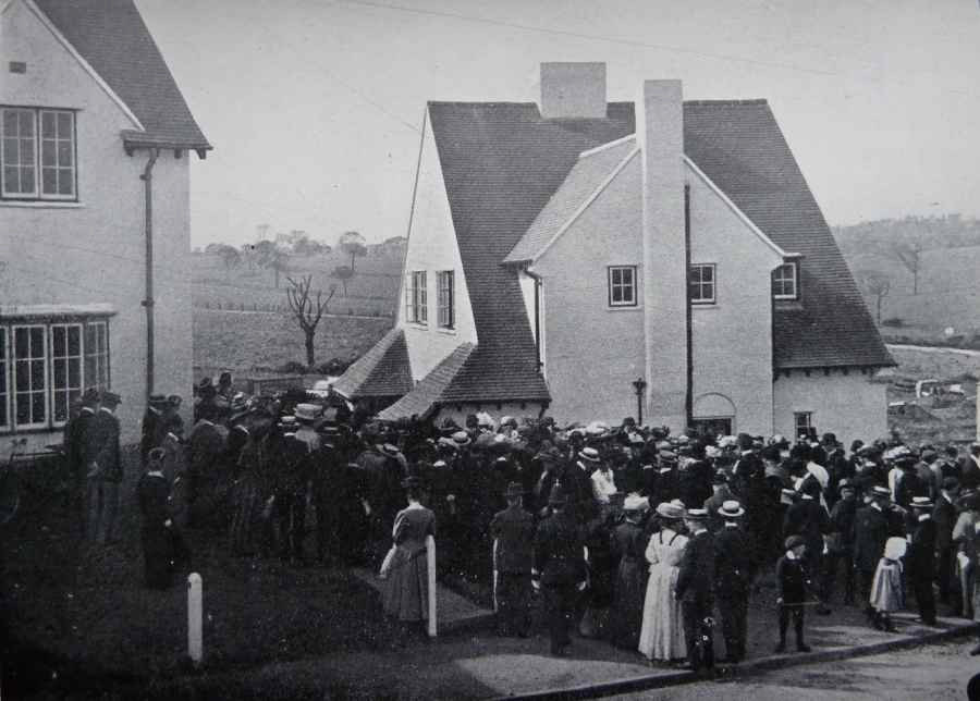 A black and white photograph showing people standing outside a house. There are fields and trees in the background.