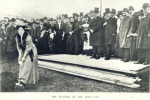 A black and white photograph showing a woman using a trowel to dig into the earth. There is a large group of people stood behind her.