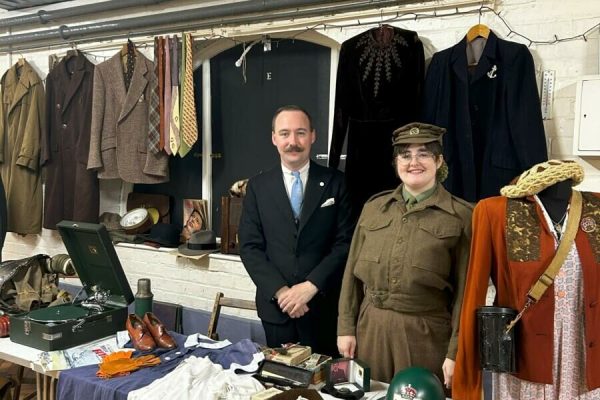Two people in historical clothes stood behind a table covered in more historical clothes